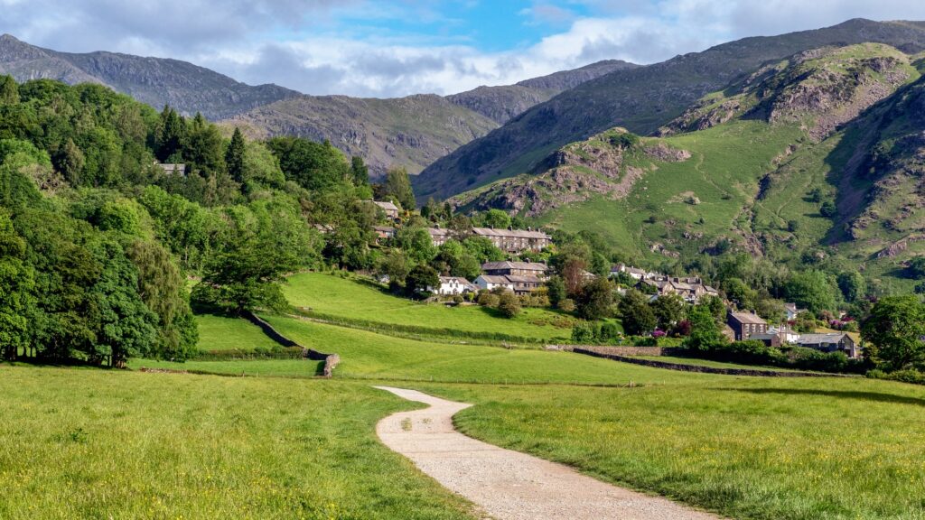 Path through a grassy field towards Coniston village with hills in the background