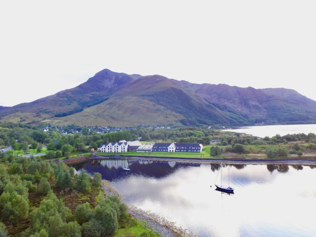 The Isles of Glencoe Hotel beside calm water with mountains behind it